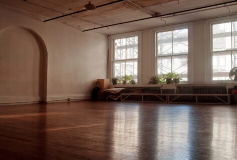 A studio with hardwood floor and arch cut out in the wall, large windows with plants placed in front let in a beam of light reflecting of the wood. Photo by Ian Douglas.