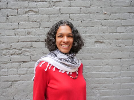 Portrait of Marýa Wethers. She has black curly hair and wears a long sleeve red top and a keffiyeh. She looks into the camera smiling brightly. Photo by Katherine Paola De La Cruz