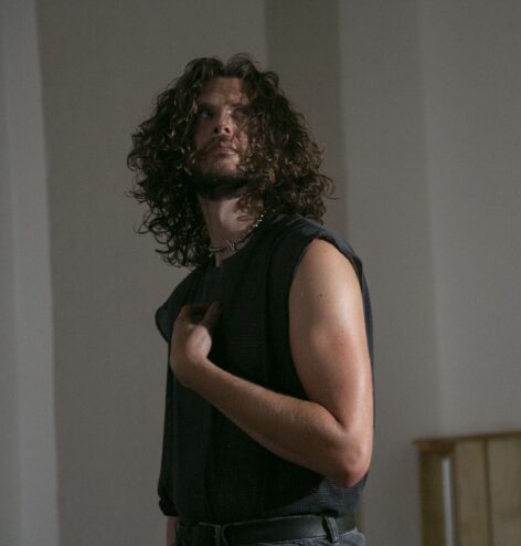 Image of a performer with brown shoulder-length curly hair and a short beard. They are cropped from the hips up and wearing a black tank top. Their upper body is pivoting front and their eyes are looking
quizzically to the upper right diagonal with their right hand pointing to their chest. Photo by Whitney Browne.