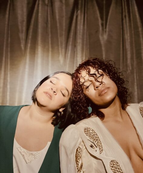Two women pictured tilting their heads inward to another with eyes closed. One is wearing a green blazer, and mover pictured to the right is wearing a white and gold embellished blazer. Photo by Tori Dominguez