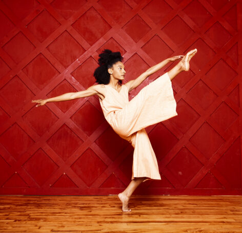 Photo of Katherine Paola De La Cruz. She balances on one leg on releve while kicking her other leg up. Her arms reach out from her torso. She has dark curly hair. Photo courtesy of the artist.