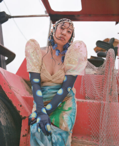 A Filipino woman in front of a red tractor draped with fish netting leans forward with arms folded over her lap, wet hair slicked around her face, and a forlorn expression. She wears a head piece with charms and jewels, blue cascading floral earrings, long polka-dotted formal gloves, and a traditional Philippine dress airbrushed with Gumby surfing a wave. Photo by Akasha Rabut.