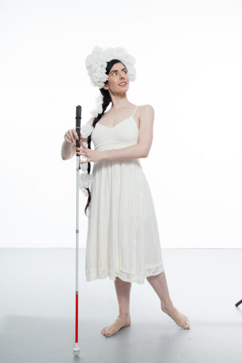 Krishna, a thin, pale skinned woman with long dark hair pulled into a braid, poses on a studio stage. She is wearing a simple white dress with her shoulders exposed and a fluffy white headdress perched on
her forehead and wrapping down her braid. She looks over her left shoulder as she lightly holds a straight cane between her hands and
points her left foot behind her. Photo Credit: Krishna Washburn & Micaela Mamede