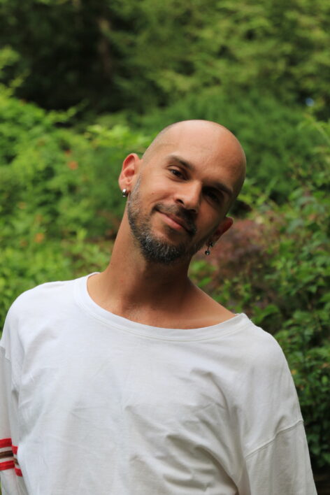 Photo portrait of Liony. Liony poses in front of greenery in a white shirt with his head tilted to the side. He has a kind smile on his face. Photo courtesy of the artist.