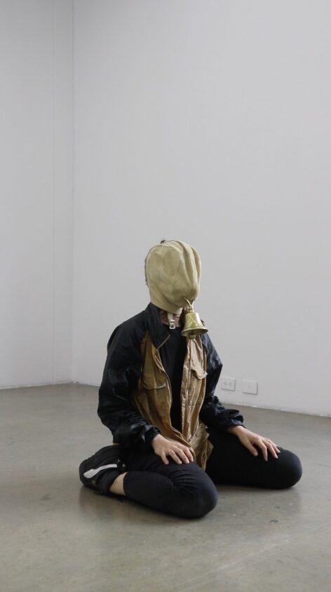 A body sits on the floor, legs bent backwards, hands resting
onto of its knees. The body is wearing black puma trainers,
black trousers, a black and gold jacket. The body wears a
mask made of suede over its face, where the mouth would
be, hangs a copper bell. The space behind the body is
painted white and the floor is painted grey. Photo courtesy of the artists.