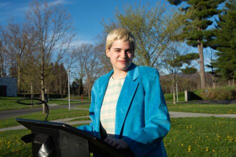Kari, a white nonbinary person with short bleached hair, stands at a
podium smiling with a teal suede blazer on. In the background, blue skies, trees and grass.