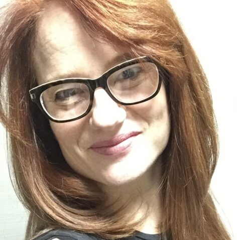 Stephanie Tack grinning in to the camera. She has dark rimmed glasses and ginger hair. Photo courtesy of the artist.