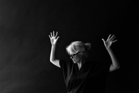 Yanira is against a black background. Her hands are raised above her head, palms forward. She looks down in profile. Photo by Simon Courchel.