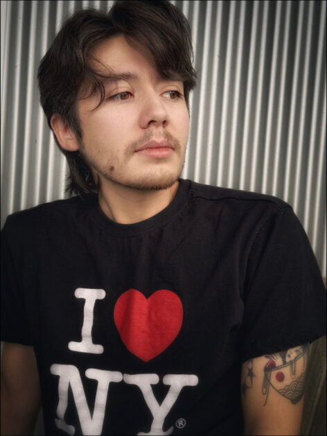 evan is pictured outside in front of a silver metal wall. The image is cropped at the chest. evan looks away from the camera and wears a black t-shirt that says 