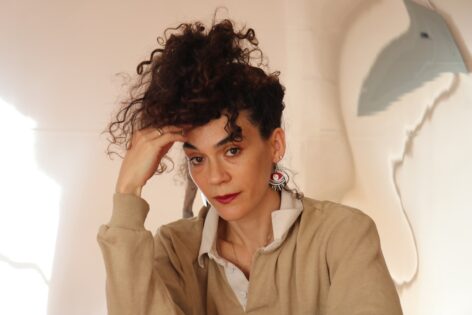 Mor Mendel gazes into the camera. Her head and hand lightly touching, hair in high ponytail. Photo by Tim Craig