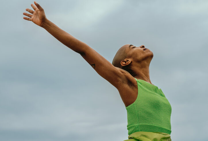 A photo of Jasmine reaching behind them in lime green. They look toward the sky with their eyes closed smiling. Photo by Jakayla Monay.