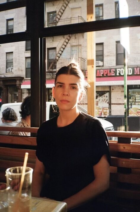 Alexa sits at a table near an open window of a restaurant; in front of her is a glass with a straw in it, she wears a black top and her hair is pulled back. Photo by Em Adamo.