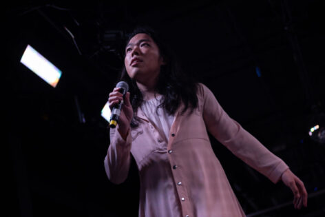 Softly holding a microphone in their hand, a young South East Asian femme person gives a longing look above the horizon.