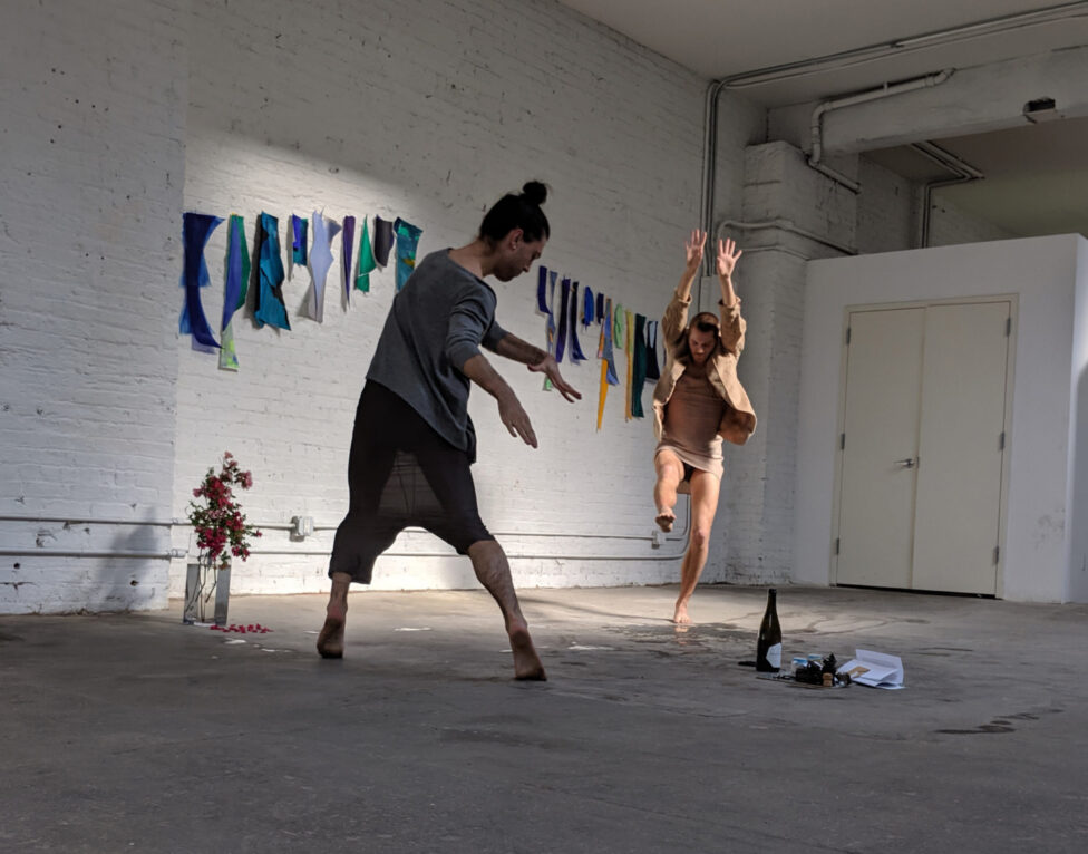 Image of two dancers moving in a white, industrial looking space. The dancer closes is moving downwards in space, the one in the background is jumping.