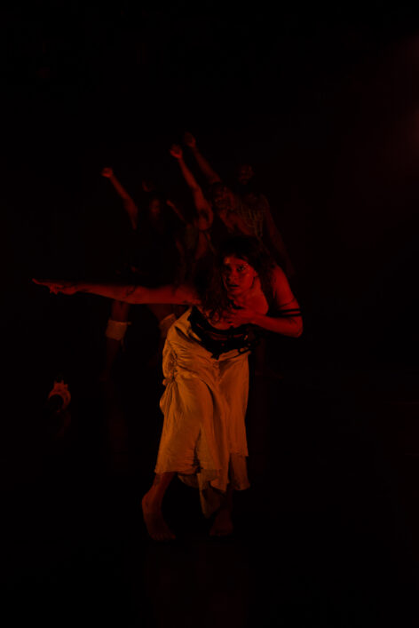 In this image Valentina stands on the foreground and they are bent over looking straight at the camera with a look that expresses fearless rage. Their left arm is extended out to the side and pointing in a sharp manner. In the background a group of dancers point at Valentina, their faces holding both shock and horror. The image is quite dark; the shadows give way to a cave like mystery. Photo by Blu.