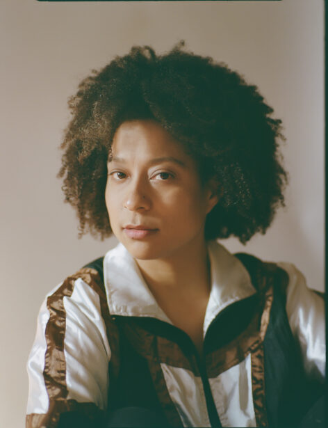 A headshot of a biracial black and white woman with a calm gaze toward the camera. She has a natural afro hairstyle and is wearing a white, black, and brown silky tracksuit jacket. Photo by Nuria Rius.