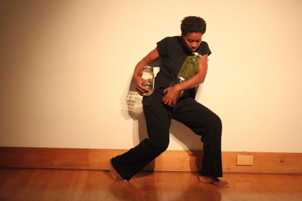 Image of a dancer dressed in all black moving, holding two glass jars in each arm. 