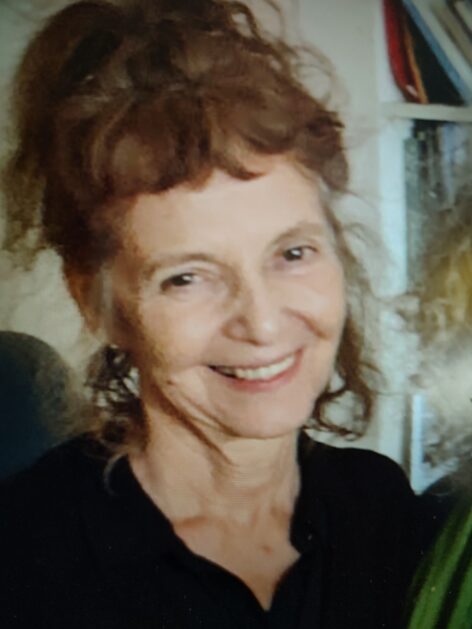 A smiling photo of Vicky wearing a black top. She is a white woman with brownish hair messily piled on top of head; her head is tilted to the right. Photo by David Tobis.