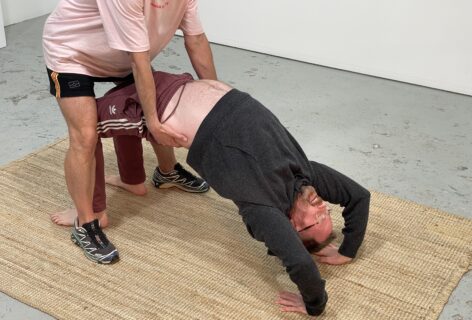 Nic Chilvers (left) helps Anthony (right) elevate his pelvis. Arching the body upward with assistance helps bring oxygen into Anthony’s lungs, while relieving fatigue and tension in the lower back, chest, and stomach. This gestural movement explores the notion of working with injury, identifying agency, and maintaining body mobility.