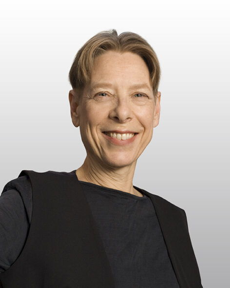 Portrait of Irene Dowd smiling into the camera. She has short blonde hair and wears a dark top with a black vest. Photo by Mathew Karas.