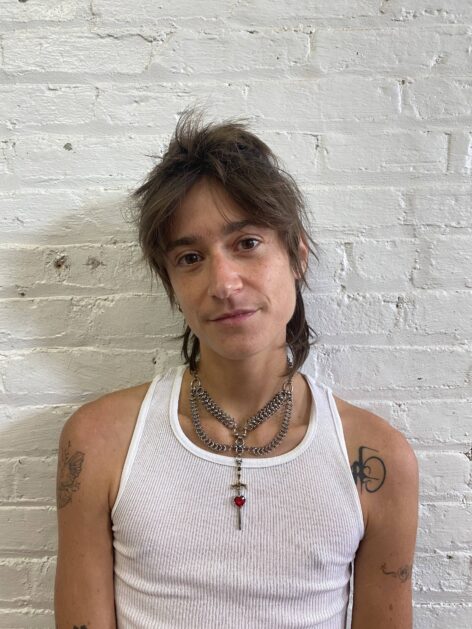 Jo is a white genderqueer person with short brown hair standing in front of a white wall wearing a white tank top. Photo courtesy of the artist.