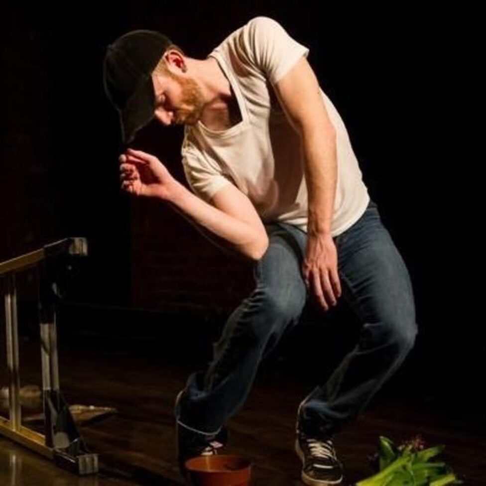 A figure in a white t-shirt and jeans leans in a crouched position, their right hand lightly holding the black baseball cap atop their head. Photo by Ian W. Douglas. 

