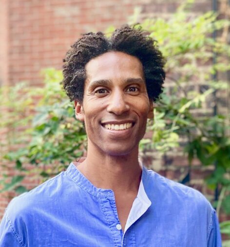 A photo of Stacy Matthew Spence smiling at the camera. He has short afro hair and wears a blue top. Photo by Michael Argilla.