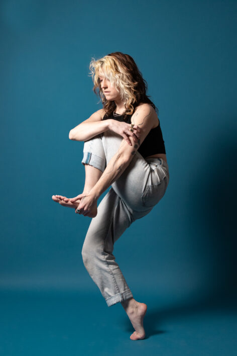 Rachel balances on the ball of one foot while holding the bottom of their other food with one hand. Their other hand is wrapped around their upper body. Their face is concentrating. Rachel wear's blue mom jeans and a black tank top and stands in front of a bright blue background. Self Portrait by Rachel Keane