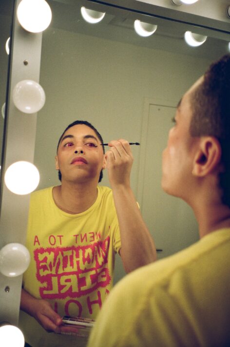 Maxi puts on eye liner in a backstage mirror with red eye contacts. She has black short hair and is slightly looking up. Photo credit Saint Pinero.