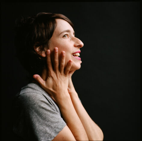 Tatyana is a light skinned, white jewish woman in profile, holding her head in her palms and looking up with a smile. Photo by Maria Baranova