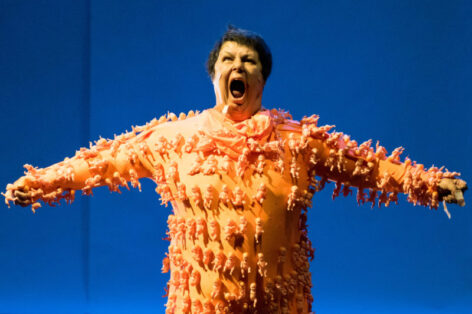 Yvonne Meier in performance. She stands with her arms spreading from her body in a t shape. She wears a peach tone onesie with small plastic babies glued on to it. Photo by Ian Douglas