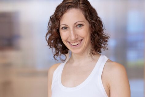 Allie is smiling warmly at the camera in front of a blurred background. She has curly hair and is wearing a white tank top. Photo courtesy of artist. Photo by Theik Smith.