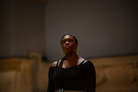 Performance photo of Ogemdi Ude, a dark skinned black woman. She wears her braided hair tied back, gold hoops and an off the shoulder black long sleeve top, she has a calm facial expression and her head is tilted up slightly and she looks beyond the camera. Photo by Rachel Keane.