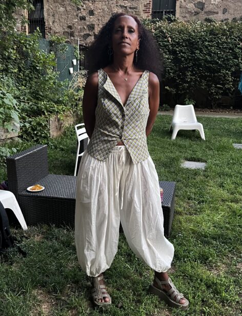 Black woman standing with hands behind her back. She is wearing a green and yellow v-neck blouse with white barrel pants. Photo by Orion Gordan.