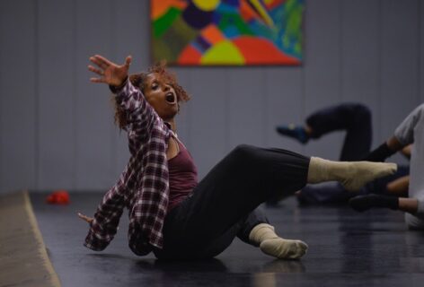 A photo of nia love seated with her arms spread wide and one leg lifted. Photo by Grant Halverson.