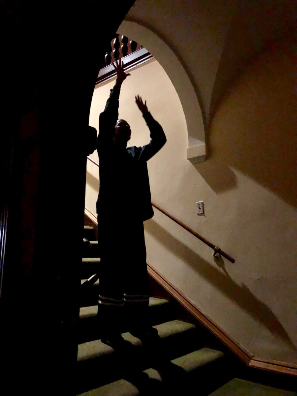 Photo of a person standing on a set of stairs, with light casting behind them, and they are lifting up their arms. 