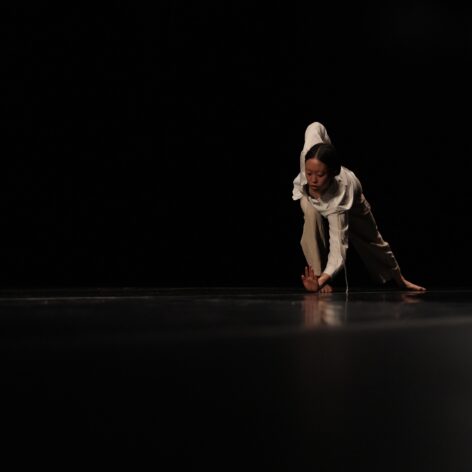 On a darkened stage Clara wears loose beige clothing while draping her torso laterally over her bent right leg as her left leg extends straight behind her. Her right arm forms an angular shape tucked behind her right side body and her gaze is fixed on her left hand’s fingertips.  Photo credit to Derek Fowles.