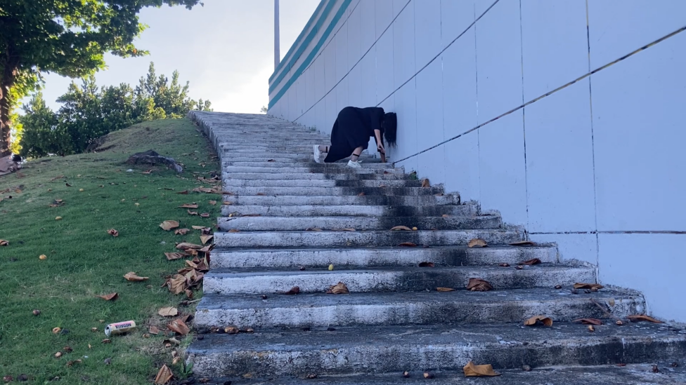 Halfway down a curved grey staircase, Nicole is in a black jumpsuit and white sneakers crouched with her head leaning against a surrounding white wall. Photo courtesy of the artist.