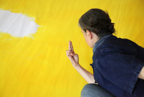 Daria holds her index and middle finger together and looks at it. We see the back of her head, her hair in a low bun while she crouches in front of a bright yellow wall. Photo courtesy of the artist.