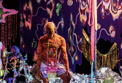 Ishmael Houston Jones stands with weight shifted into one hip with his gaze looking downwards. In the background a maximalist design with purple pink yellow and blue design. Ishmael wears a long sleeve orange top and a light colored pair of shorts. There is a small pink plastic purse hanging from his neck. Photo of Robbie Sweeney.