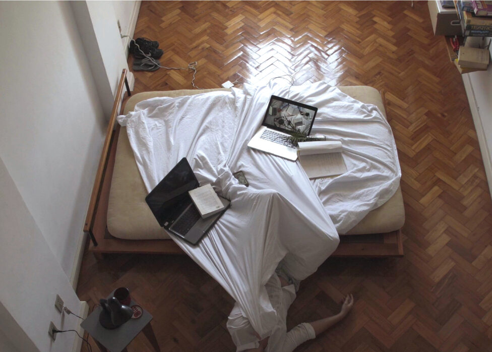 A bed with white sheets and a wooden frame sit in a room with miscellaneous objects around the perimeter. The bottom half of Sofia's body lays halfway on the bed and the top half on the wooden floor. Two laptops and a couple of notebooks remain on the bed's white sheets. Photo courtesy of the artist.