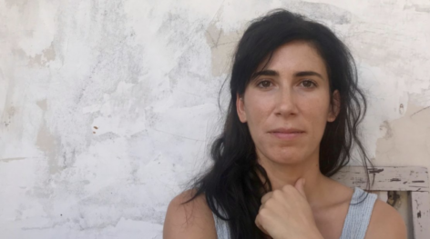 A woman with olive skin and billowing dark hair stares directly forward, touching her throat