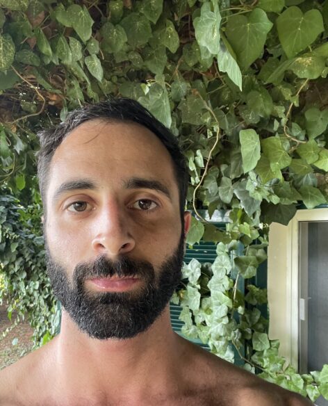 Portrait of Charlie Prince. He has short black hair, a black beard. He is looking at the camera with a neutral expression. He is surrounded by green vines. The photo is taken in Deir el Qamar, Lebannon. Photo courtesy of the artist.