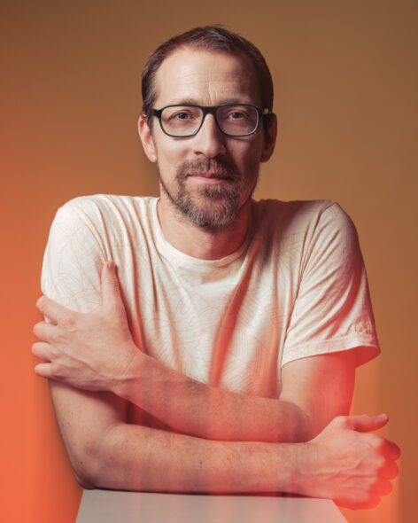 Portrait of Gergely Talló. He is wrapping his arms around his torso. He wears a white t-short and dark rectangle frame glasses. He looks straight into the camera. Photo by Domolky Daniel.