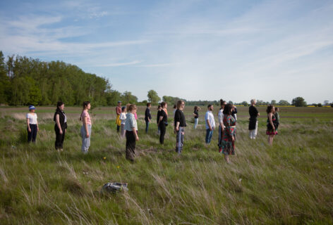 In a field of lush green grass. People stand facing east all gazing off into the distance. The sky opens up above them in its blue clear splendor. Photo courtesy of Ewan McLaren.