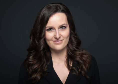 Headshot of Brigitta Kovács. She looks straight into the camera with a grin. She has long dark brown hair and wears a black v neck top. Photo by Dániel Dömölk.