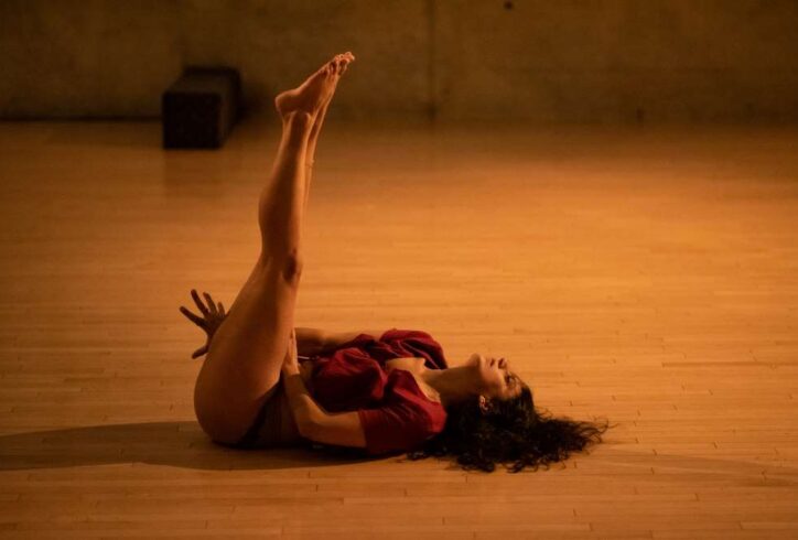 Sahar Damoni lies on the hardwood floor at the Judson Memorial church. She wears a red dress and has her legs up in the air while one hand reaches in between and the other on her thigh. Her eyes are closed. Photo by Ian Douglas