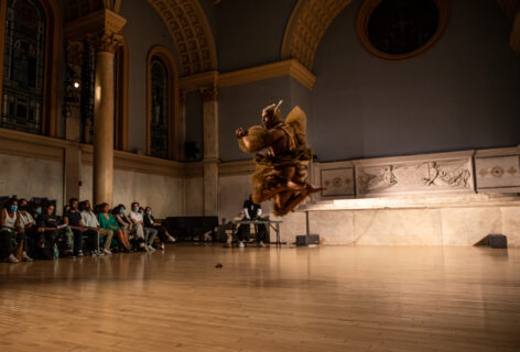 Photo of Maxi Hawkeye Canion. They are performing for Movement Research at the Judson Church. They wear a light brown costume made of many layers of tulle and a head piece with two long ears sticking straight up. They are in mid air, jumping, knees bent. In the background the audience looks on.