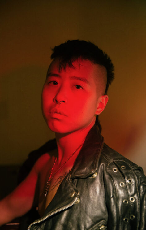 With short black hair shaved on the sides, Riven looks into the camera as he is lit by a red light and draped with a black leather jacket.
credits: Mengwen Cao