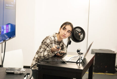 Leonor leans over a desk with a remote in her hand. She sits in front of a laptop in a plaid top photo by Abigail Montes